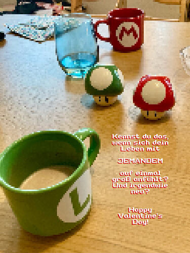 Alt-Text: Zwei Salzstreuer in Form eines roten Super-Pilzes und eines grünen Extra-Leben-Pilzes stehen auf einem Holztisch, daneben eine grüne Luigi- und eine rote Mario-Tasse. Daneben der Text: Kennst du das, wenn sich dein Leben mit JEMANDEM auf einmal groß anfühlt? Und irgendwie neu? Happy Valentine’s Day!